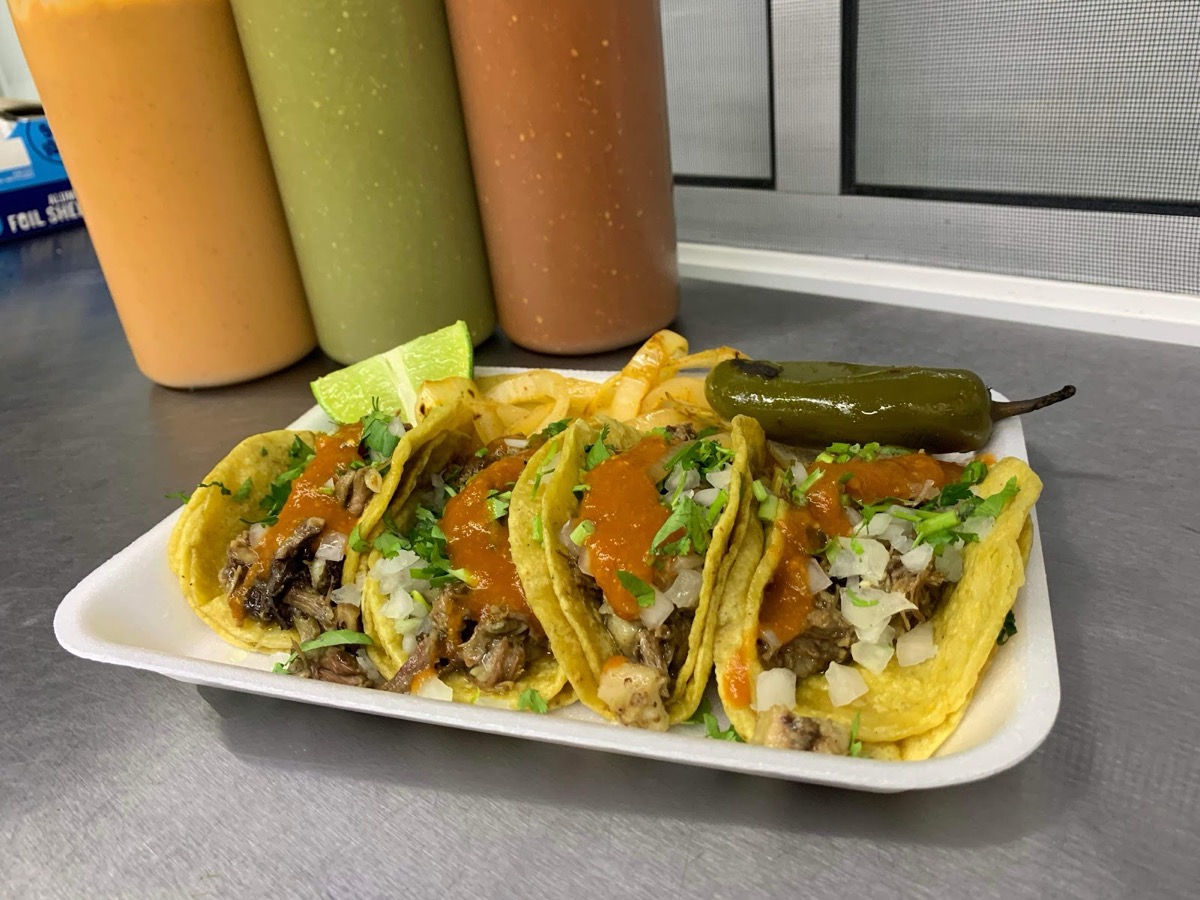Four tacos on a tray with fries, a lime wedge and a grilled jalapeño, salsa bottles behind